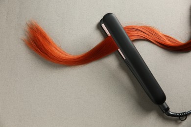 Photo of Modern straightener with red hair lock on grey background, top view