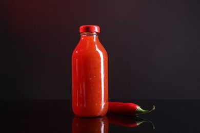 Photo of Spicy chili sauce in bottle and pepper against dark background