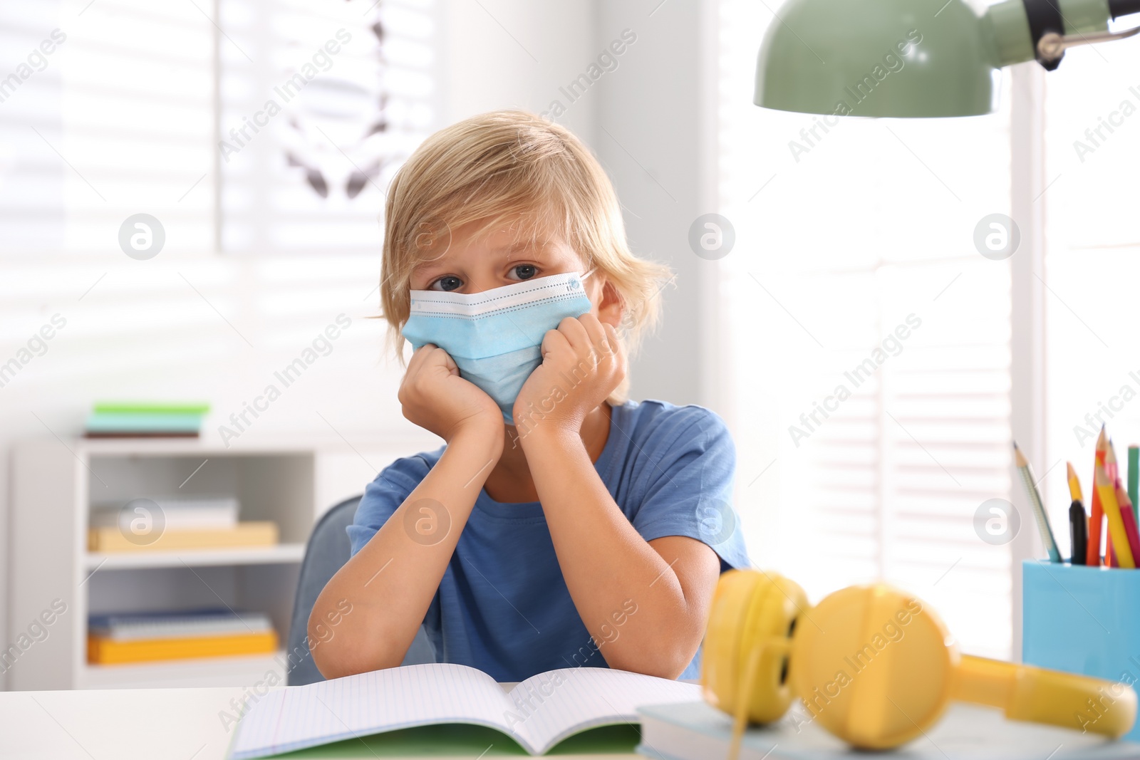 Photo of Sad little boy in mask with exercise book at home. Distance learning during COVID-19 pandemic