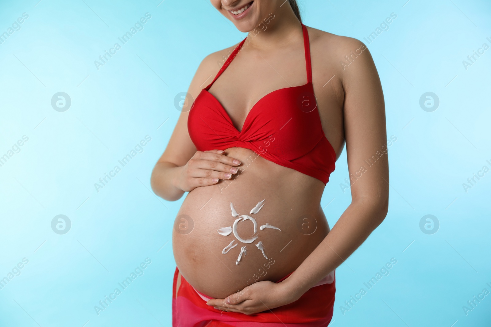 Photo of Young pregnant woman with sun protection cream on belly against light blue background, closeup