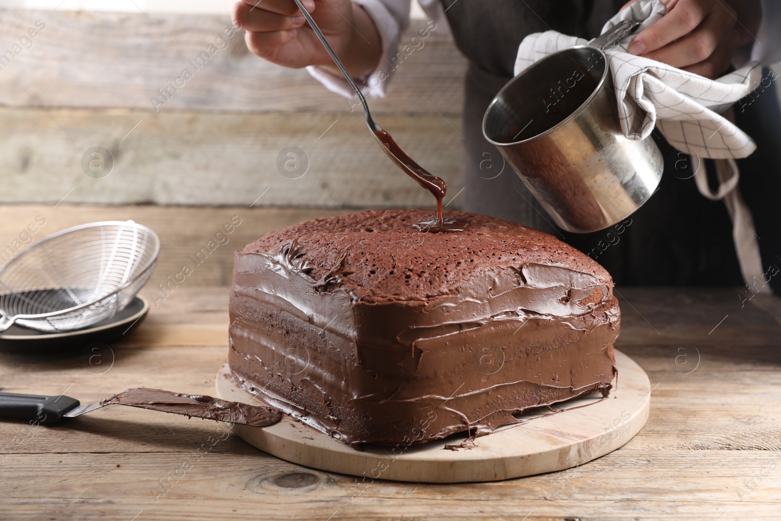 Photo of Woman pouring chocolate cream into homemade sponge cake at wooden table, closeup