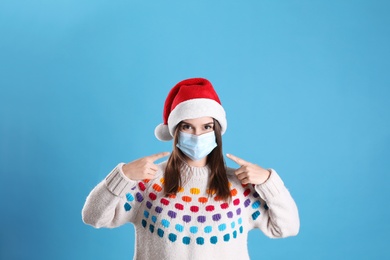 Photo of Pretty woman in Santa hat and medical mask on light blue background
