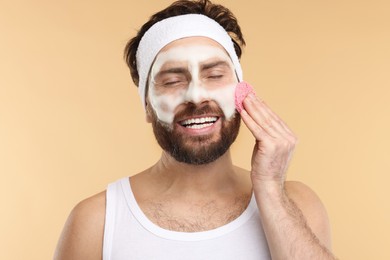 Photo of Man with headband washing his face using sponge on beige background