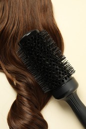 Professional round brush with brown hair strand on beige background, top view