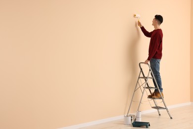 Photo of Young man painting wall with roller on stepladder indoors, space for text. Room renovation