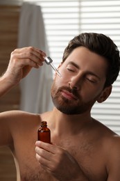 Handsome man applying cosmetic serum onto his face indoors