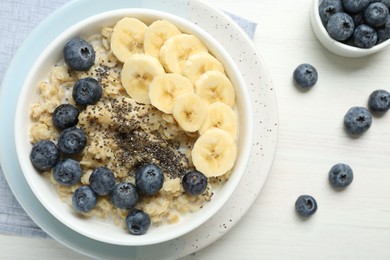 Tasty oatmeal with banana, blueberries and chia seeds served in bowl on white wooden table, flat lay