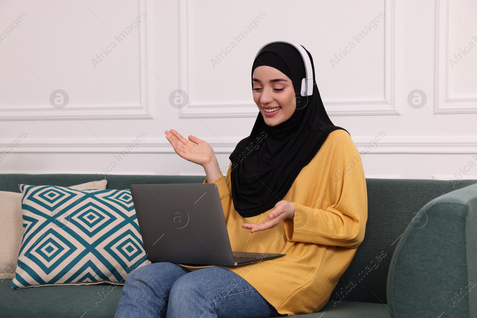 Photo of Muslim woman in hijab and headphones using video chat on laptop indoors