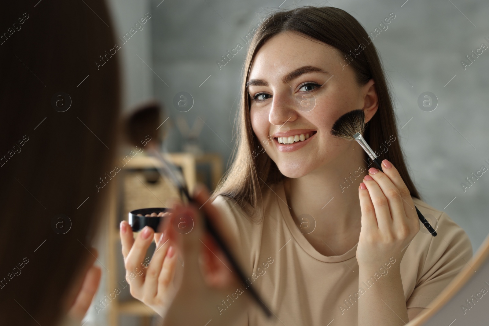 Photo of Smiling woman with freckles applying makeup near mirror indoors