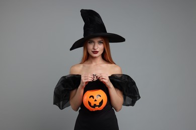 Photo of Young woman in scary witch costume with pumpkin bucket on light grey background. Halloween celebration