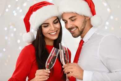 Photo of Happy young couple in Santa hats with glasses of champagne against blurred festive lights. Christmas celebration