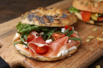 Photo of Tasty bagel with cured ham, cream cheese, tomatoes and arugula on wooden board, closeup