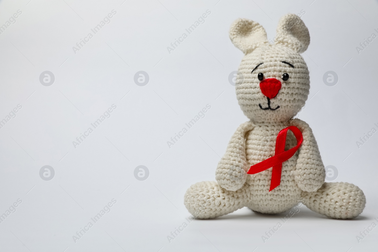 Photo of Cute knitted toy bunny with red ribbon on light grey background, space for text. AIDS disease awareness