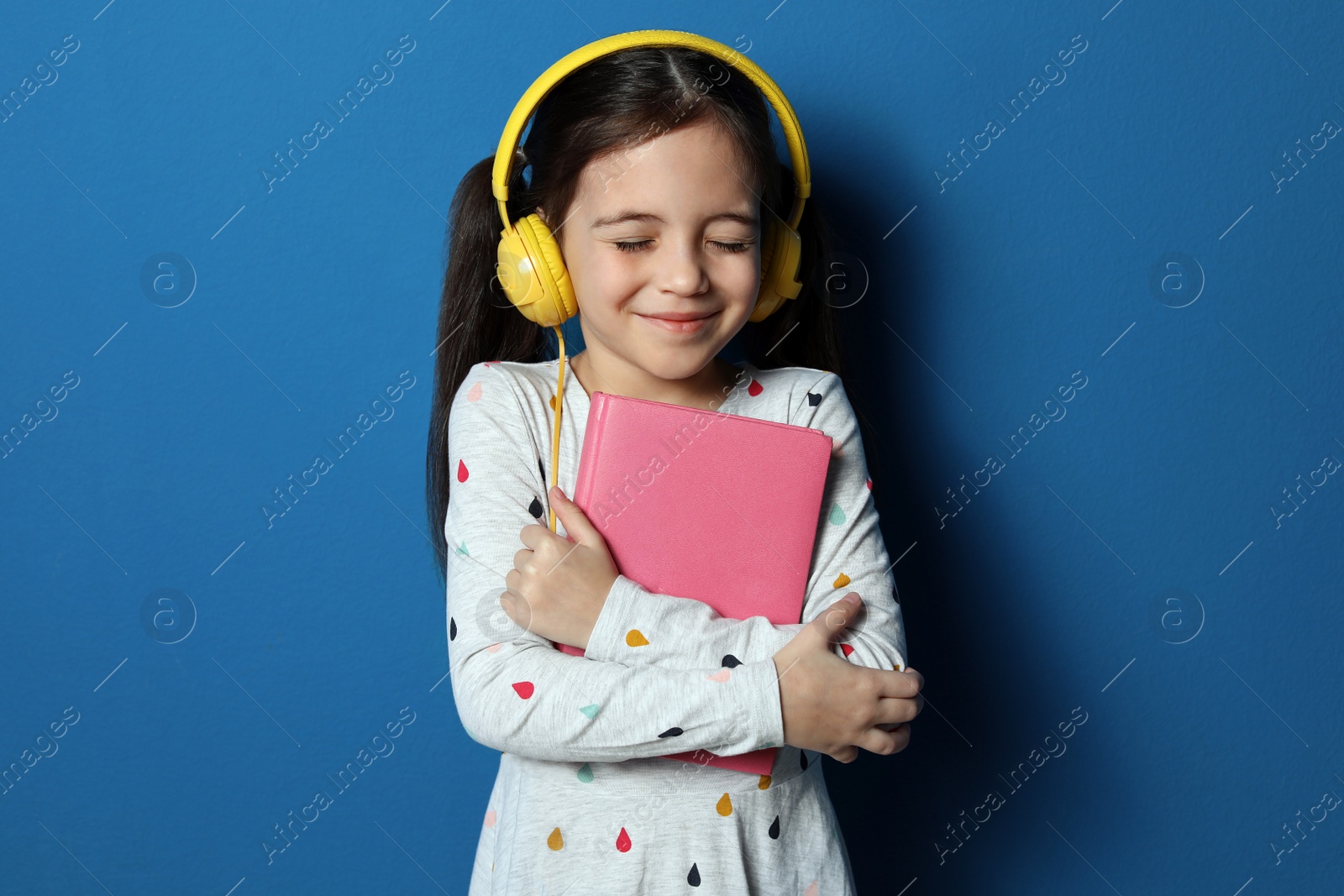 Photo of Cute little girl with headphones listening to audiobook on blue background