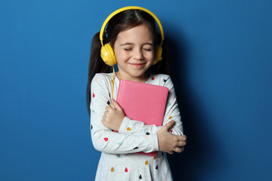 Cute little girl with headphones listening to audiobook on blue background