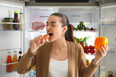 Photo of Young woman eating sausage near open refrigerator