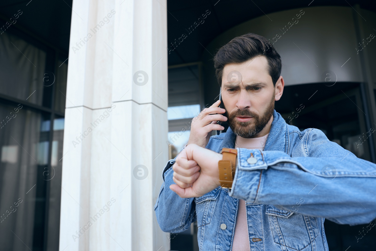 Photo of Emotional man talking on smartphone and looking at watch near building. Being late concept