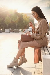 Photo of Fashionable young woman with stylish bag on bench outdoors