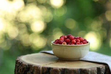 Bowl of tasty wild strawberries on wooden stump against blurred green background. Space for text