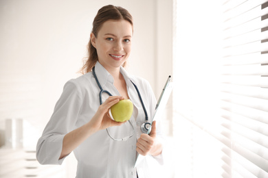Photo of Nutritionist with apple and clipboard near window in office. Space for text