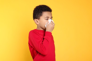 African-American boy blowing nose in tissue on yellow background, space for text. Cold symptoms