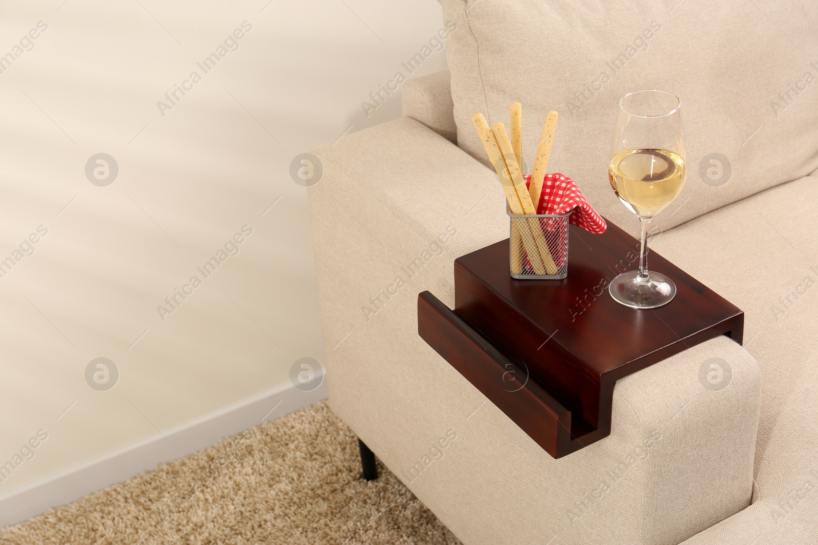 Photo of Glass of white wine and breadsticks on sofa with wooden armrest table in room, space for text. Interior element