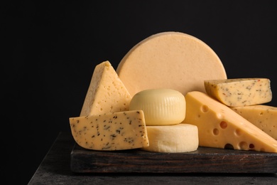 Wooden board with different sorts of cheese on table against black background, space for text