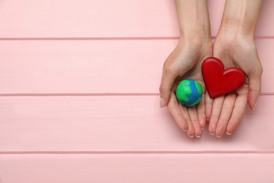 Photo of Happy Earth Day. Woman holding plasticine planet and decorative heart at pink wooden table, top view with space for text