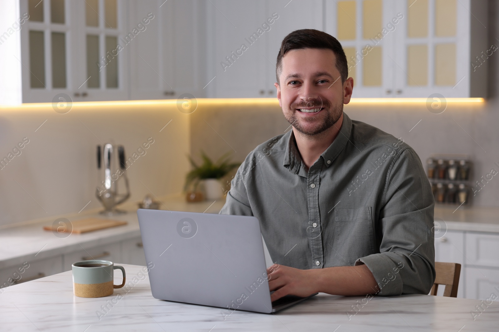 Photo of Happy man working on laptop at white marble table in kitchen
