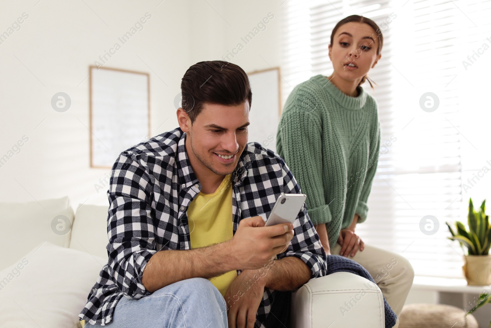 Photo of Distrustful woman peering into boyfriend's smartphone at home. Jealousy in relationship