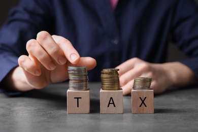 Photo of Woman with word Tax made of wooden cubes and coins at grey table, closeup