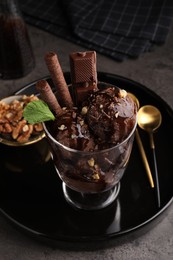 Photo of Tasty chocolate ice cream with sauce, nuts and wafer rolls in glass dessert bowl on dark grey table