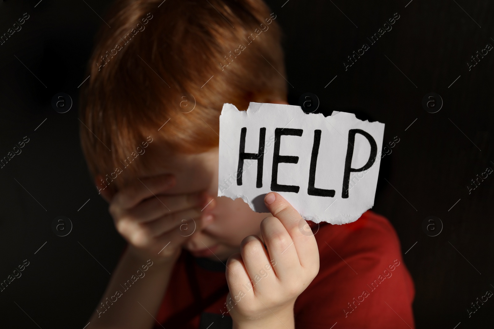 Photo of Little boy holding piecepaper with word Help against black background, focus on hand. Domestic violence concept