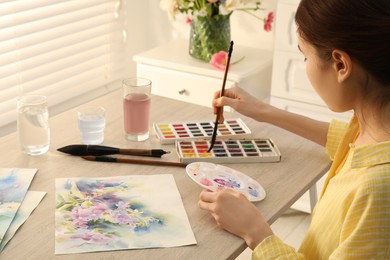 Photo of Woman painting flowers with watercolor at white wooden table indoors. Creative artwork