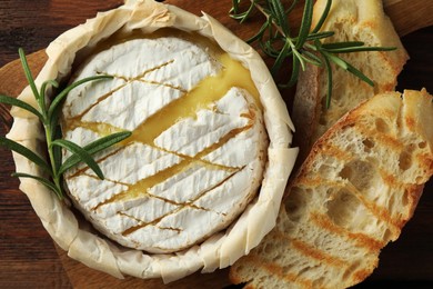 Photo of Tasty baked brie cheese, bread and rosemary on wooden table, top view