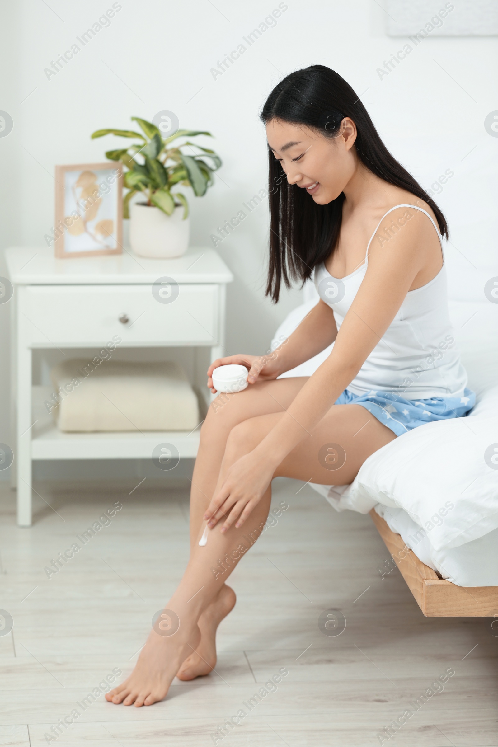 Photo of Beautiful young Asian woman applying body cream on leg in bedroom, space for text