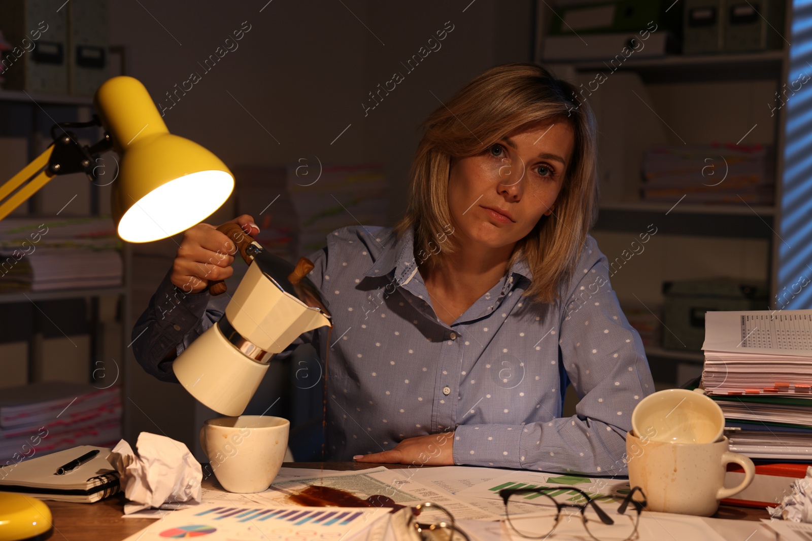 Photo of Overwhelmed woman pouring coffee past cup at workplace in office at night