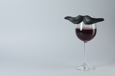 Glass of red wine with funny fake mustache on grey background, space for text