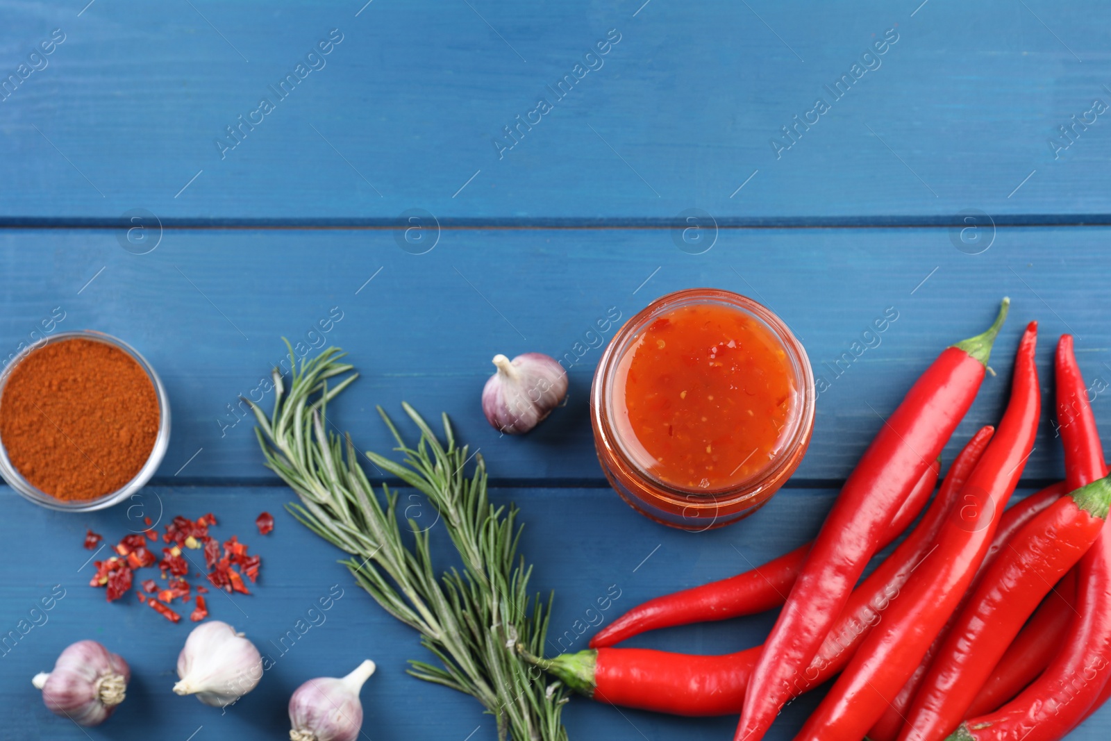 Photo of Spicy chili sauce in jar and ingredients on blue wooden table, flat lay. Space for text