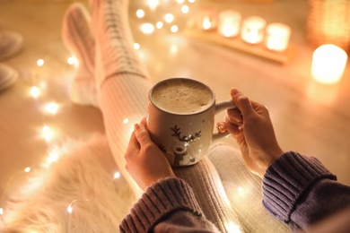 Photo of Woman with cup of drink on floor against blurred Christmas lights, closeup