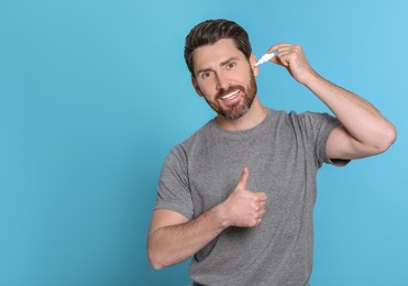 Man using ear drops and showing thumbs up on light blue background, space for text