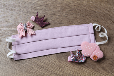 Photo of Homemade protective mask and adorable girly hair clips on wooden background, flat lay. Sewing for child