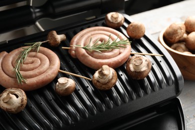 Photo of Electric grill with homemade sausages, rosemary and mushrooms on rustic wooden table, closeup