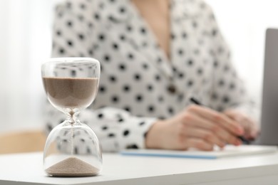 Hourglass with flowing sand on desk. Woman taking notes indoors, selective focus