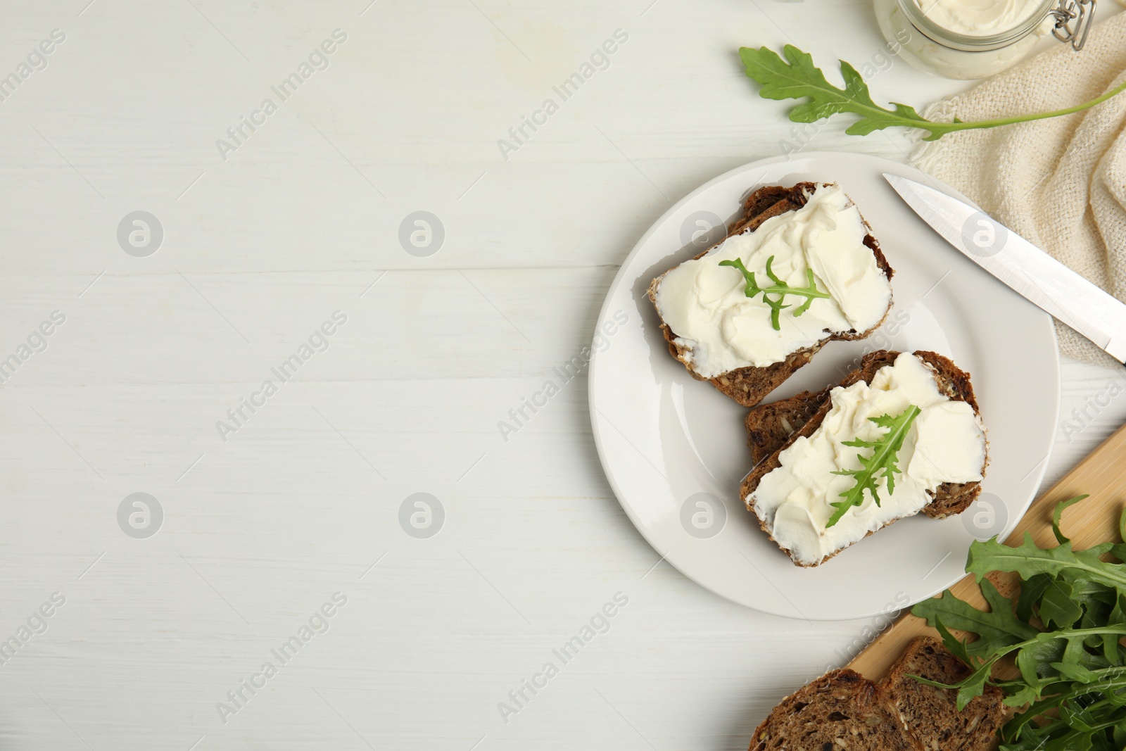 Photo of Bread with cream cheese and arugula on white wooden table, flat lay. Space for text