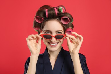 Photo of Beautiful young woman in silk bathrobe with hair curlers and sunglasses on red background