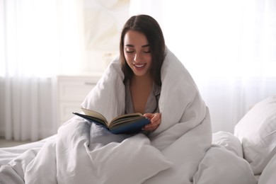 Photo of Beautiful young woman wrapped with soft blanket reading book on bed at home