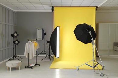 Photo of Yellow photo background, rack with clothes and professional lighting equipment in modern studio