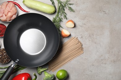 Empty iron wok surrounded by raw ingredients on grey table, flat lay. Space for text