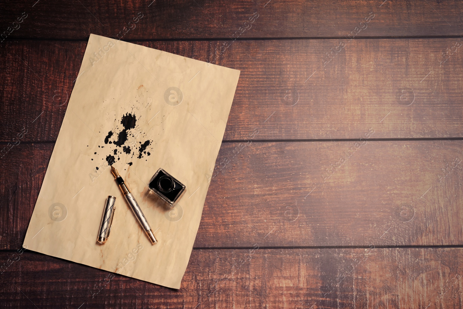 Photo of Fountain pen, inkwell and vintage parchment with ink stains on wooden table, top view. Space for text